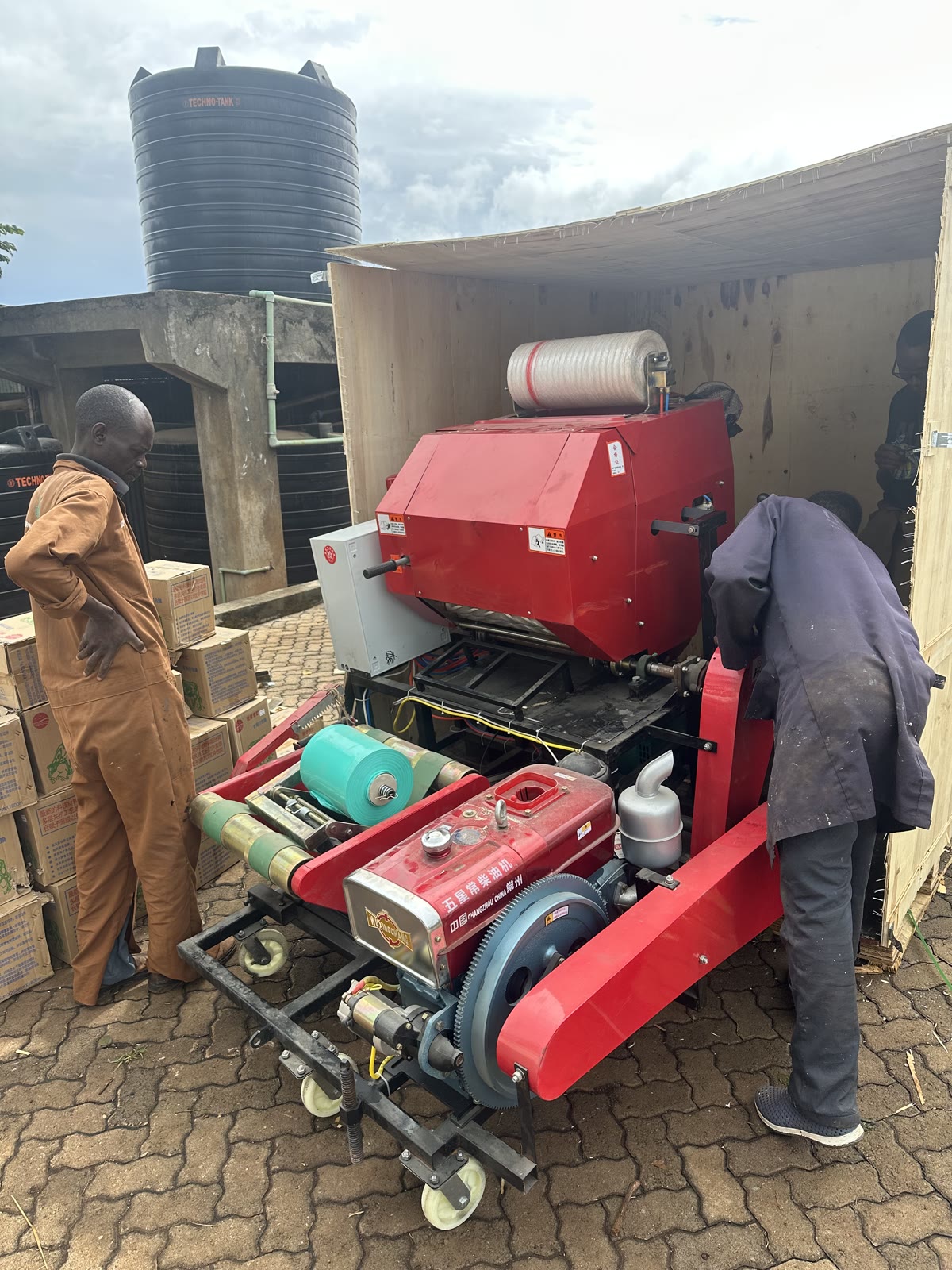 Two workers moving a red diesel silage baler wrapper out of its timber shipping crate at a farm in Kiambu County